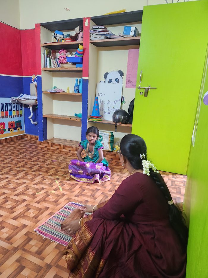 A young child seated on a mat facing a woman in a maroon saree, at the start of classes in August 2025