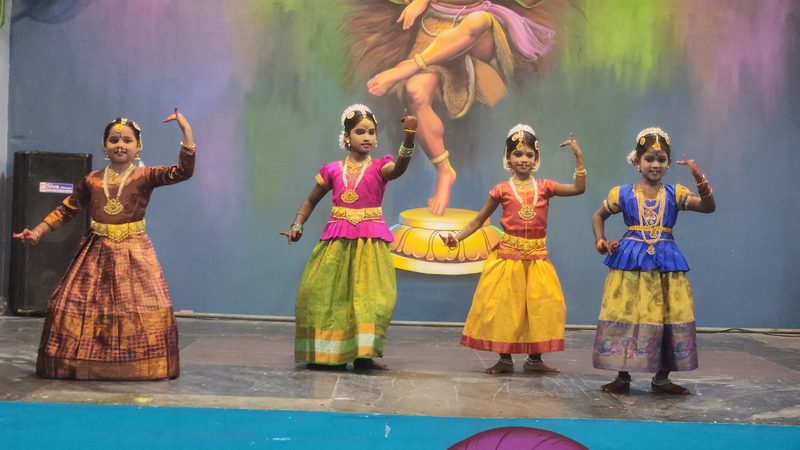 Four young students in Bharatanatyam costume performing on stage in front of a Nataraja backdrop