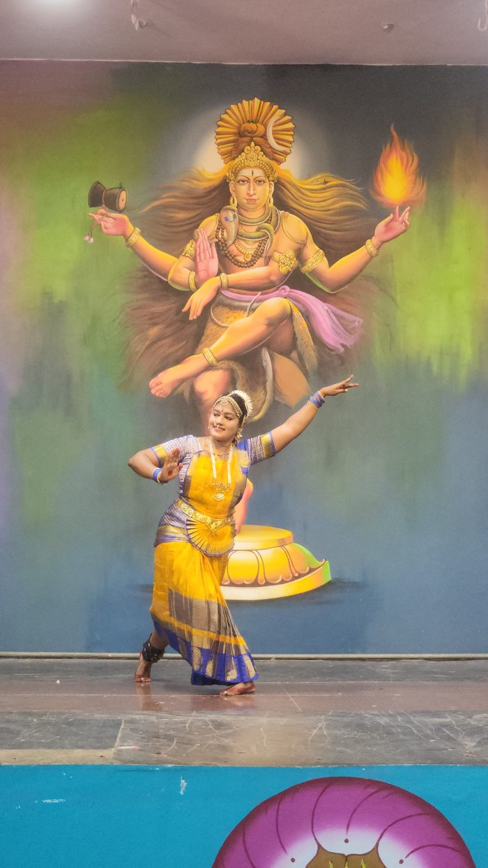 A young student in a yellow Bharatanatyam costume performing a solo on stage
